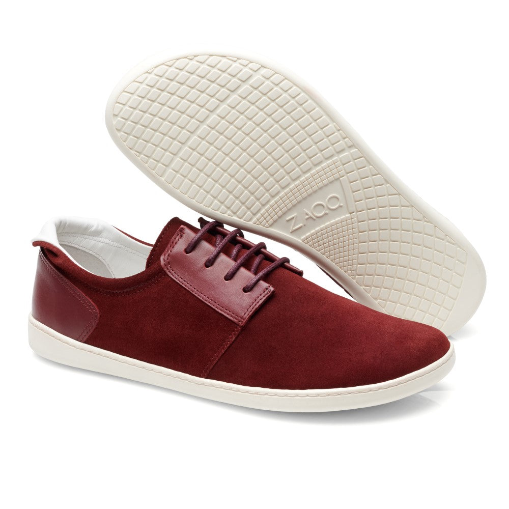 Ein Paar PIQUANT Bordeaux Leder-Sneaker mit flexibler weißer Sohle. Ein Schuh steht aufrecht und zeigt die Seite und die geschnürte Vorderseite, während der andere so positioniert ist, dass die strukturierte Sohle sichtbar ist. Der Markenname ZAQQ ist auf diesen stylischen Barfuß-Sneakern sichtbar.