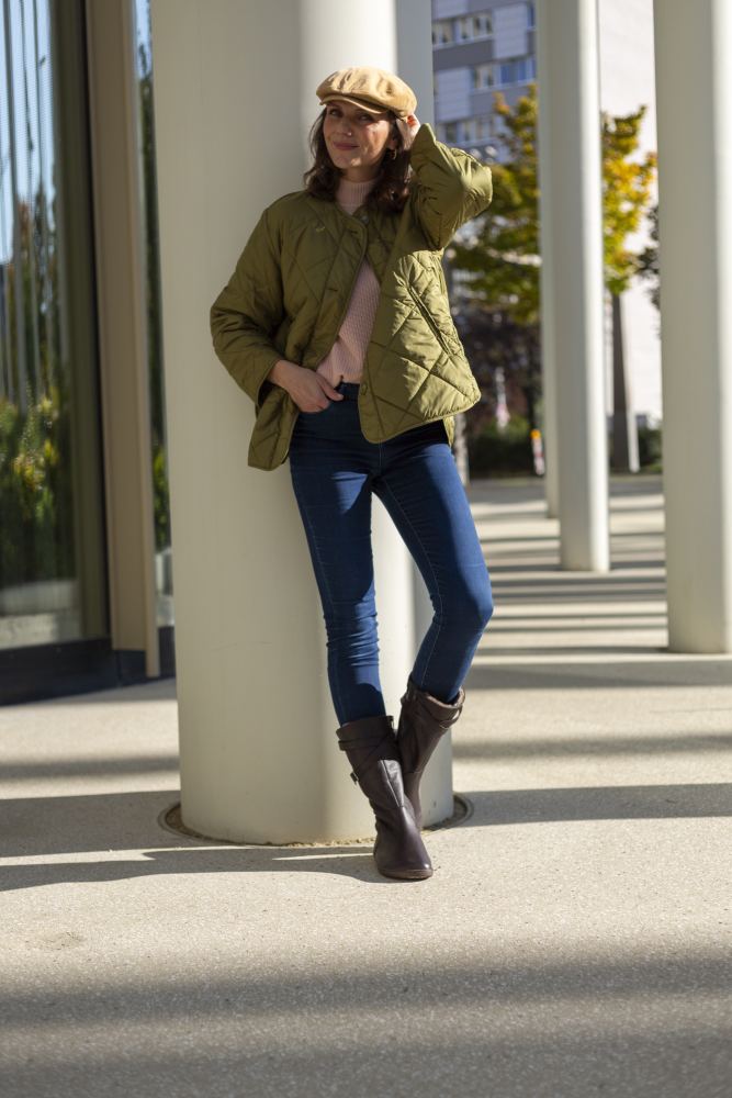 Im Freien lehnt eine Person an einer weißen Säule und betont Tragekomfort mit ihrer beigen Mütze, der grünen Steppjacke, dem rosa Pullover, den blauen Jeans und dem braunen ATTRAQT Winter Brown von ZAQQ. Sonnenlicht wirft lange Schatten auf den Bürgersteig, während Bäume in herbstlichen Farben eine malerische Kulisse bilden.