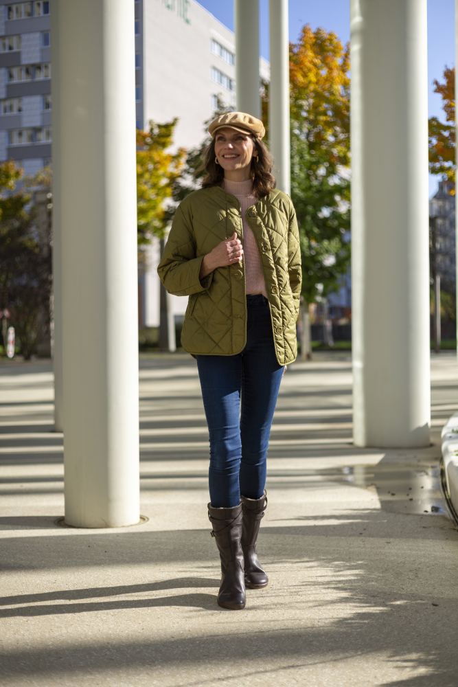 Eine Frau in einer grünen Steppjacke, Jeans und ZAQQ ATTRAQT Winter Brown-Stiefeln geht selbstbewusst unter hohen weißen Säulen hindurch. Sie lächelt und trägt eine hellbraune Schiebermütze. Im Hintergrund sind Gebäude und Herbstbäume unter einem klaren blauen Himmel zu sehen, der mit jedem Schritt Wärme und Geborgenheit ausstrahlt.