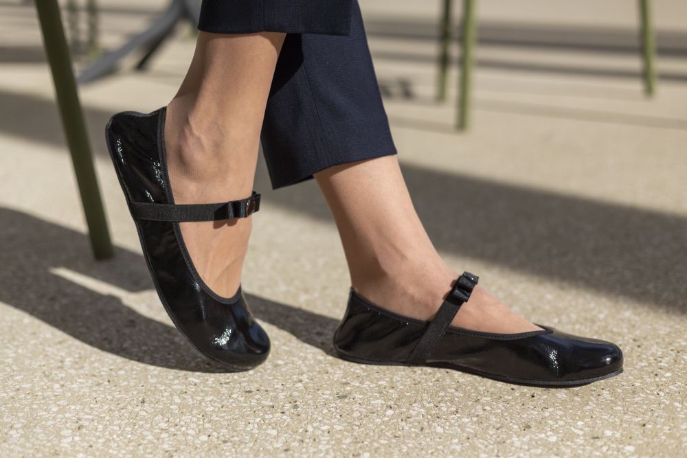 Nahaufnahme der Beine einer Person, die schwarze, glänzende TWIST CASEMIRO-Schuhe von ZAQQ mit einer kleinen Schleife an jedem Schuh trägt. Sie sitzt auf einer strukturierten Oberfläche und trägt dunkle Hosen. Schattenlinien fallen elegant auf den Boden im Hintergrund und ergänzen die flexible Sohle des Schuhs für ultimativen Komfort.