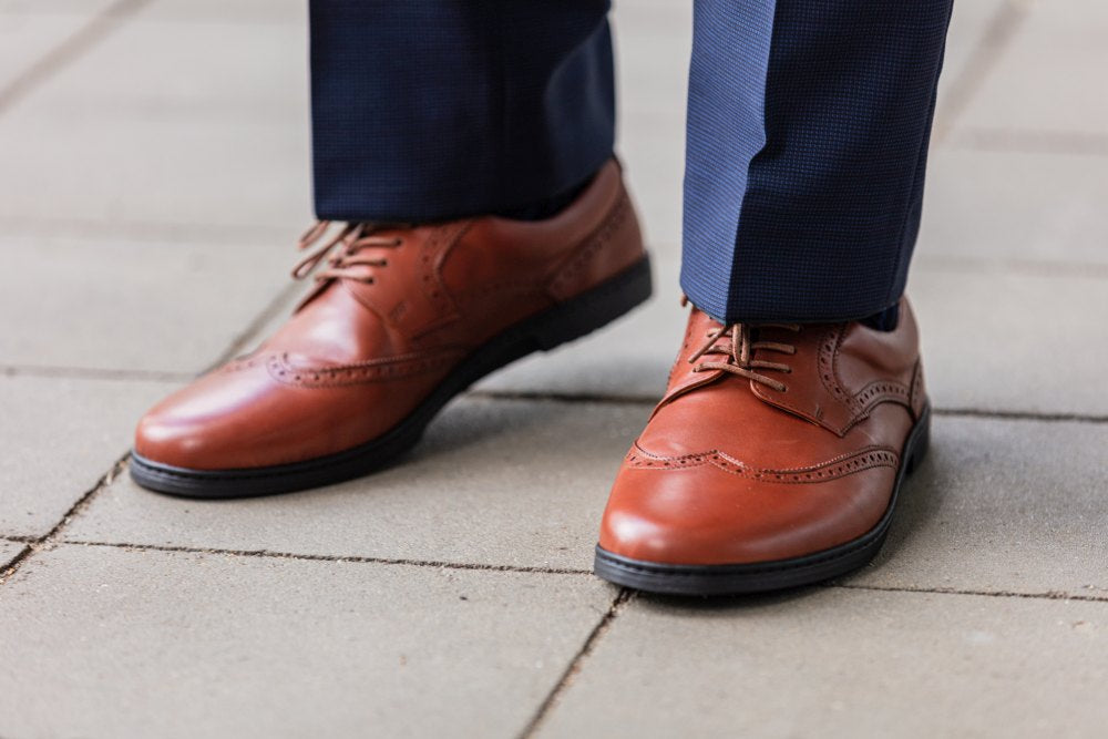 Jemand, der BRIQ Brogue Cognac-Schuhe von ZAQQ und eine marineblaue Hose trägt, steht auf einem gefliesten Boden.