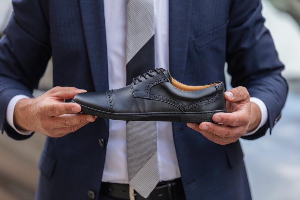 Eine Person in marineblauem Anzug und gestreifter Krawatte hält den edlen BRIQ Captoe Black von ZAQQ und präsentiert seine Brogue-Details. Der Fokus auf den Schuh unterstreicht die Eleganz dieses raffinierten Businessschuhs vor einem unscharfen Hintergrund.