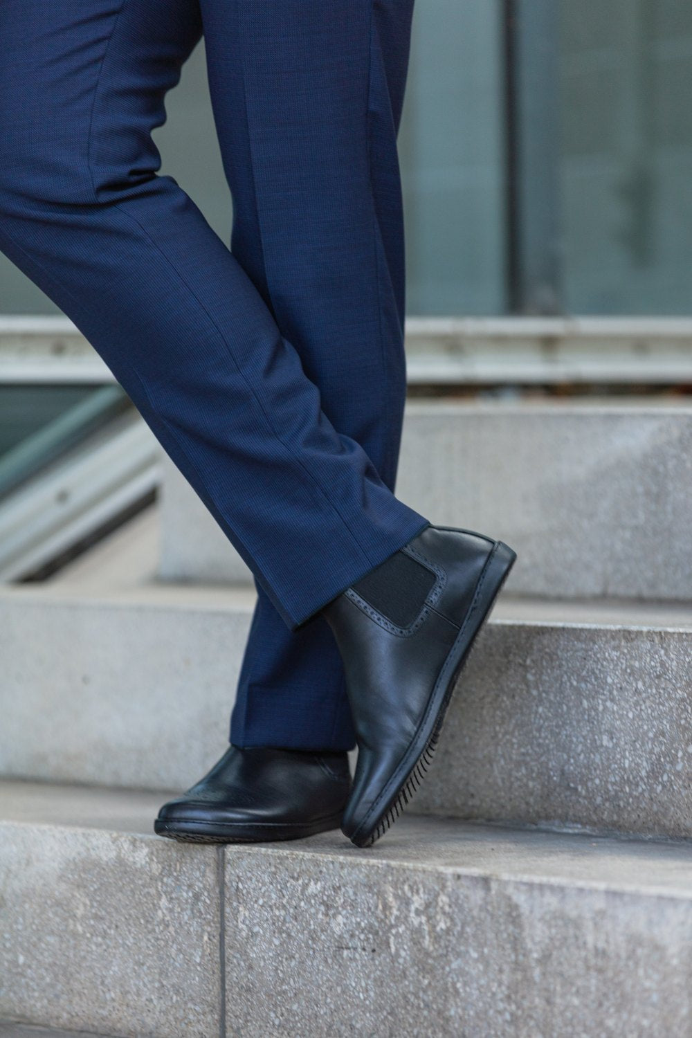 Nahaufnahme einer Person, die eine blaue Hose und schwarze EQUITY Brogue-Schuhe von ZAQQ aus Premium Nappaleder trägt. Sie steht auf Steinstufen und im Hintergrund ist ein modernes Gebäude zu sehen.