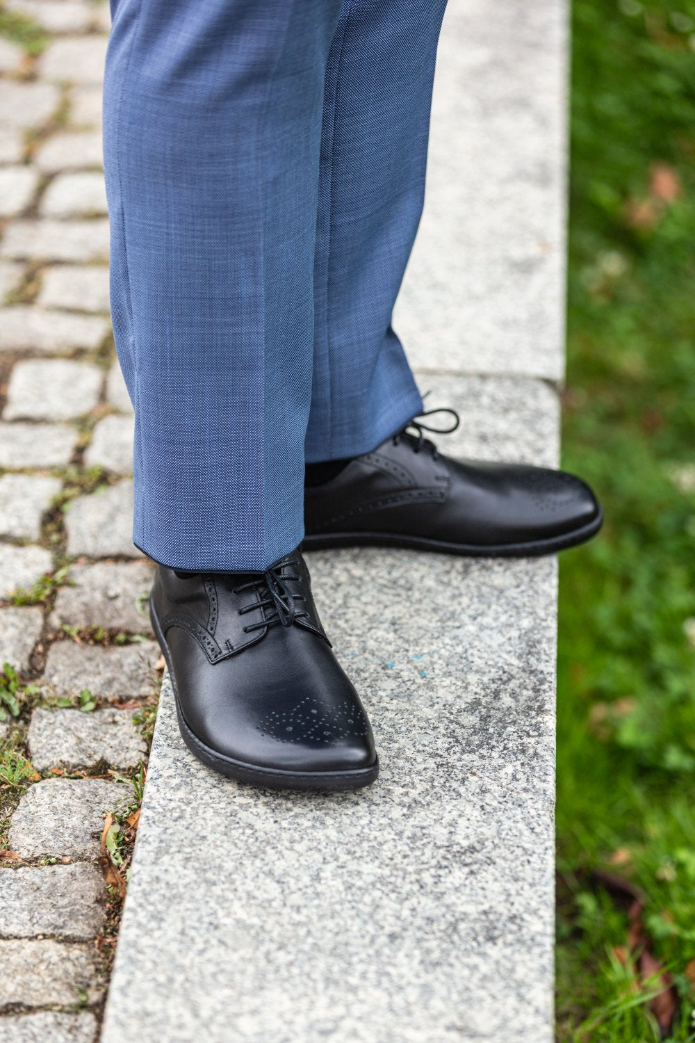 Eine Person, die die stylischen schwarzen ZAQQ PEAQ Brogue Schuhe und eine blaue Hose trägt, steht auf einem Steinweg, wo Kopfsteinpflaster mit Gras verschmilzt, was auf eine formelle oder halbformelle Außenumgebung hindeutet.