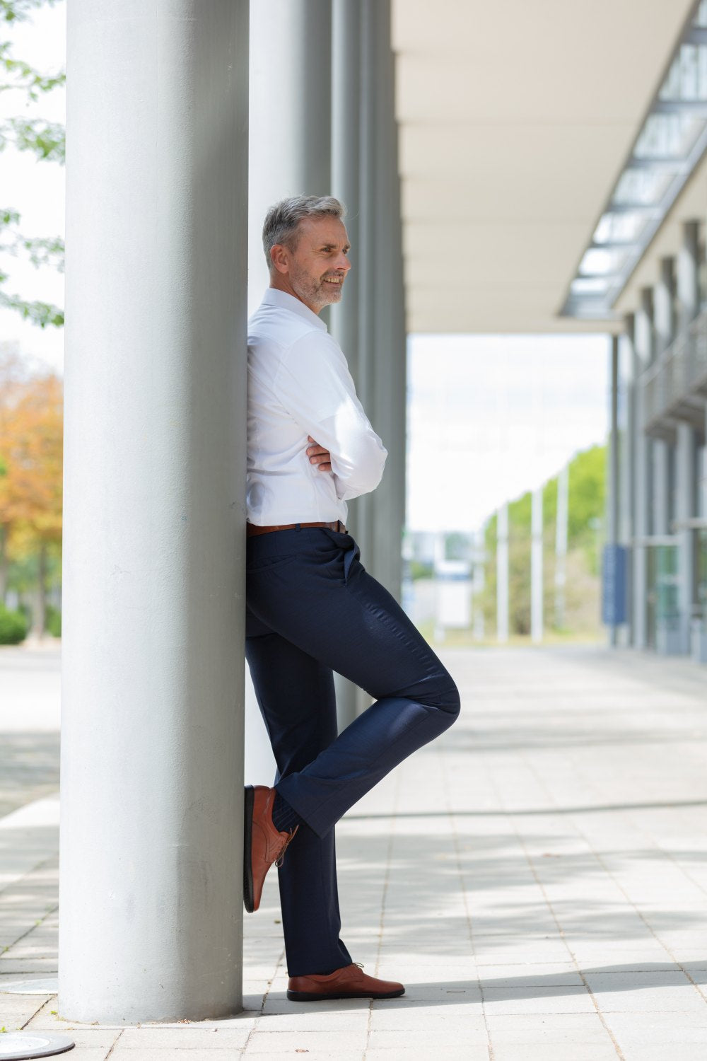 Ein Mann im Business-Outfit lehnt mit verschränkten Armen an einer hohen, grauen Säule im Freien. Mit nachdenklichem Gesichtsausdruck blickt er zur Seite. Seine PEAQ Cognac-Schuhe von ZAQQ glitzern im Sonnenlicht, während er auf einem von Säulen gesäumten Gehweg steht und auf das moderne Gebäude vor ihm blickt.