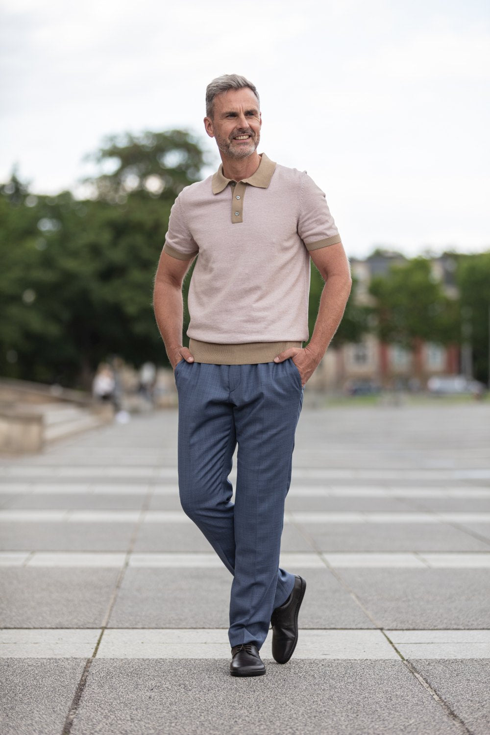 Ein Mann mit kurzen grauen Haaren läuft selbstbewusst in einem hellbeigen Poloshirt und einer blauen Hose durch die Gegend und verkörpert damit Business-Casual-Chic. Seine Hände sind in den Taschen, er trägt elegante dunkelbraune PEAQ-Schuhe von ZAQQ und lächelt an einem klaren Tag vor einer Kulisse aus Bäumen und entfernten Gebäuden.