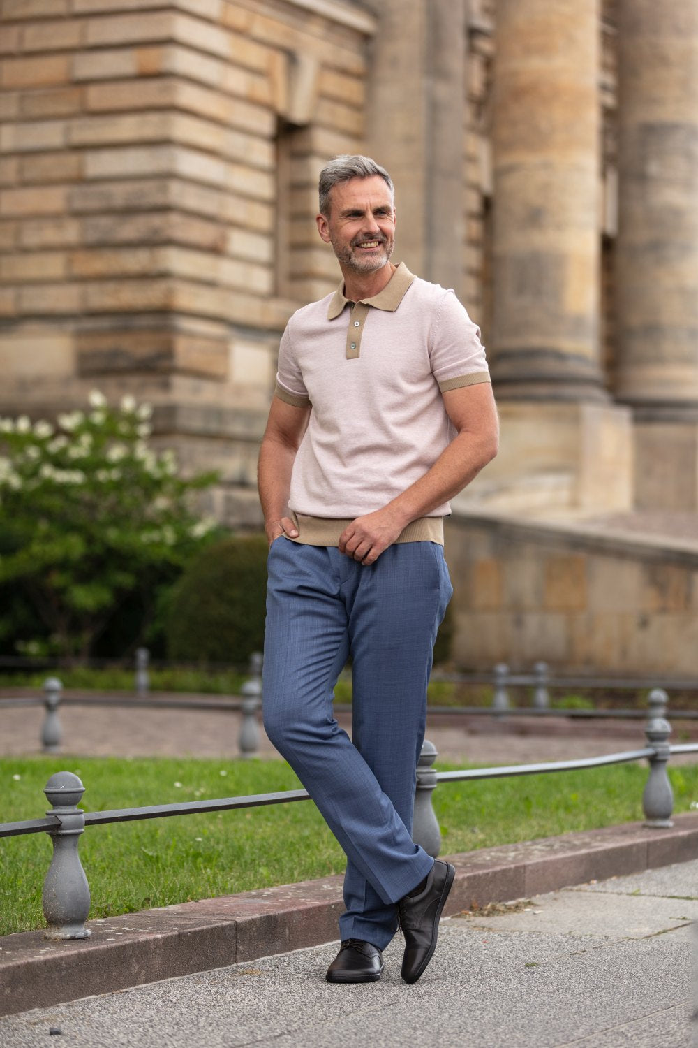 Ein Mann mit grauem Haar und Bart steht lächelnd draußen und verströmt eine lässig-business-mäßige Ausstrahlung. Er trägt ein helles Poloshirt, blaue Hosen und elegante PEAQ Dark Brown-Schuhe von ZAQQ aus feinem Nappaleder. Er steht vor einem Gebäude mit großen Säulen und lehnt an einem Metallgeländer.