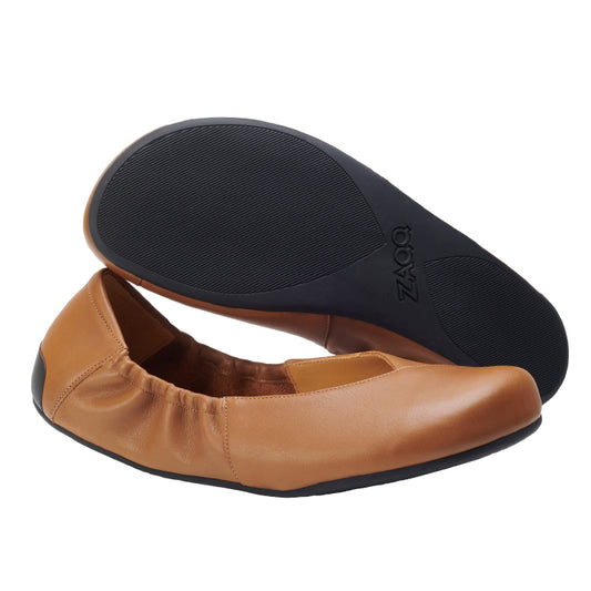 Präsentiert werden hellbraune Lederballerinas mit schwarzen Gummisohlen. Ein Schuh liegt flach und zeigt die flexible Sohle, während der andere aufrecht steht und sein schlankes Design und die elastischen Einsätze zur Schau stellt. Erleben Sie die Eleganz von ZAQQs MAIQA Brown bei jedem Schritt.