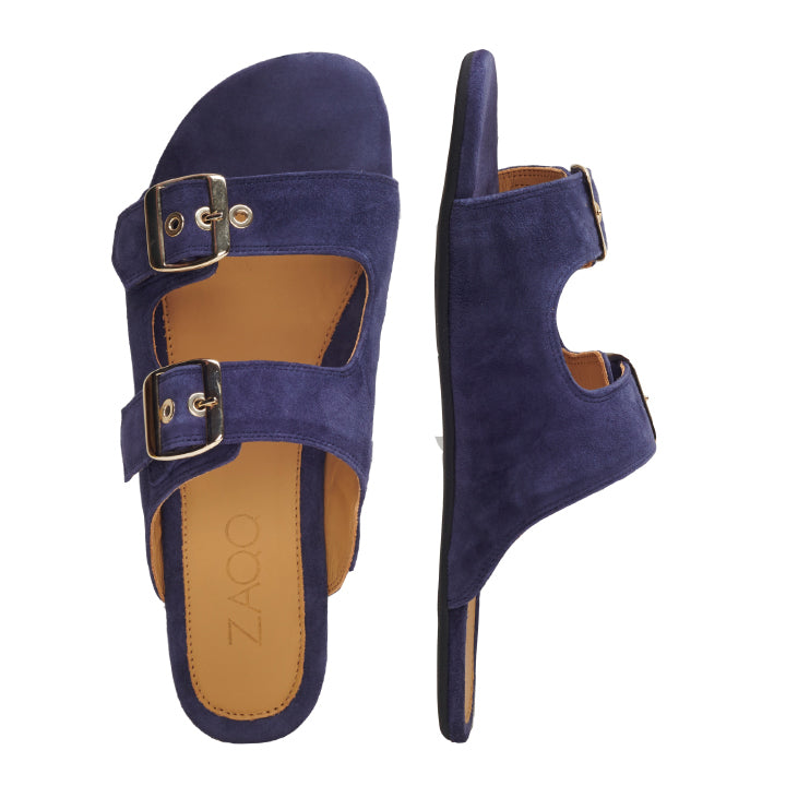 Ein Paar ZAQQ UQO Blue Sandalen, gefertigt aus marineblauem Veloursleder, mit zwei verstellbaren Riemen mit goldenen Schnallen. Sowohl in der Draufsicht als auch in der Seitenansicht dargestellt, liegt eine Sandale flach, während die andere aufrecht steht und ihr Design elegant zur Schau stellt.