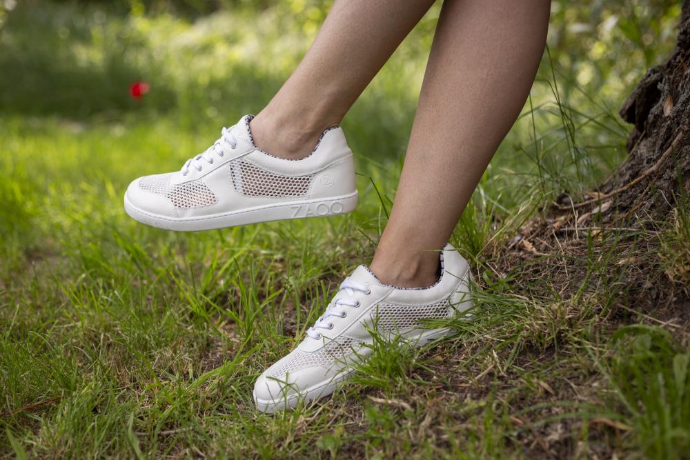 Eine Nahaufnahme zeigt eine Person, die auf Gras steht und die weißen ALIQANTE-Sneaker von ZAQQ trägt. Diese Schuhe zeichnen sich durch robuste Sohlen und ein gewebtes Design aus, das für hervorragende Atmungsaktivität sorgt. Im Hintergrund unterstreichen grüne Blätter und eine kleine rote Blume diese ruhige Umgebung.