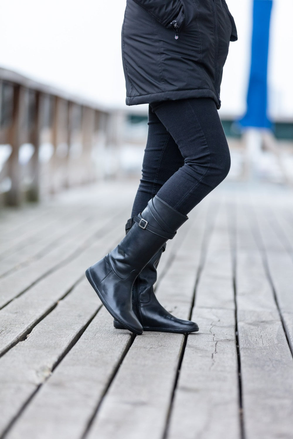 Eine Person in schwarzem Mantel, Jeans und ZAQQs ATTRAQT Nappa Black Boots steht auf einem Holzsteg. Das Bild hebt die untere Körperhälfte mit einem wunderschön verschwommenen Hintergrund hervor.