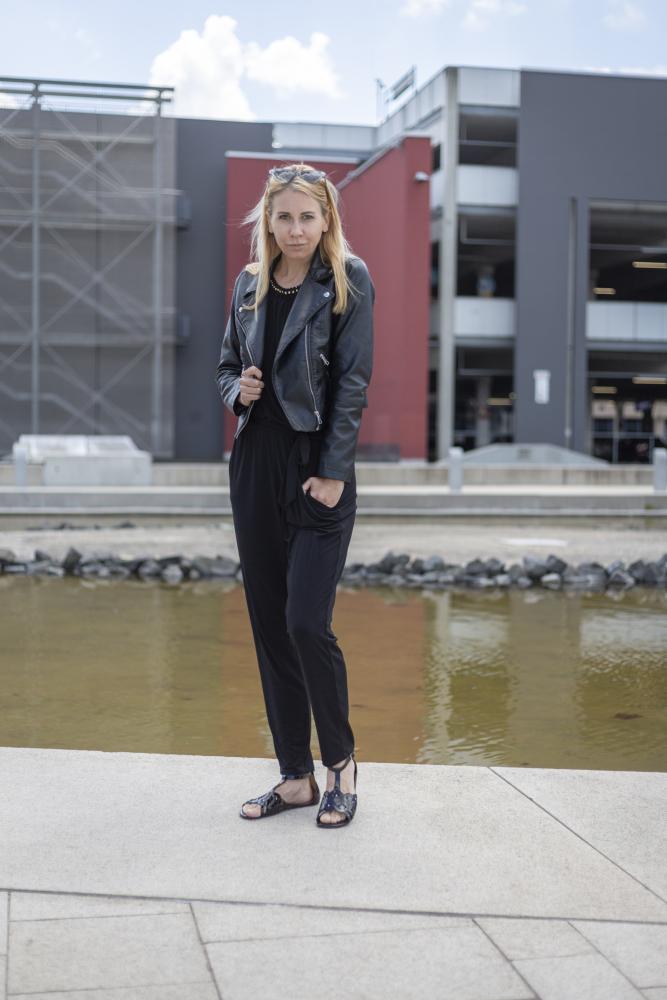 Eine Frau steht selbstbewusst neben einem spiegelnden Wasserspiel und trägt eine schwarze BOUQET Jacke und einen Jumpsuit von ZAQQ. Auf ihrem Kopf ruht eine stylische Sonnenbrille, während sie ihren eleganten Look mit modischen Barfußsandalen vervollständigt, die vor der Kulisse moderner Gebäude sowohl Stil als auch Rutschfestigkeit bieten.