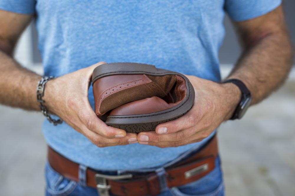 Eine Person in blauem Hemd und Jeans hält einen gefalteten ZAQQ BRIQ Brogue Antique Cognac Schuh mit flexibler Sohle und präsentiert sein biegsames Design aus hochwertigem Kalbsleder.