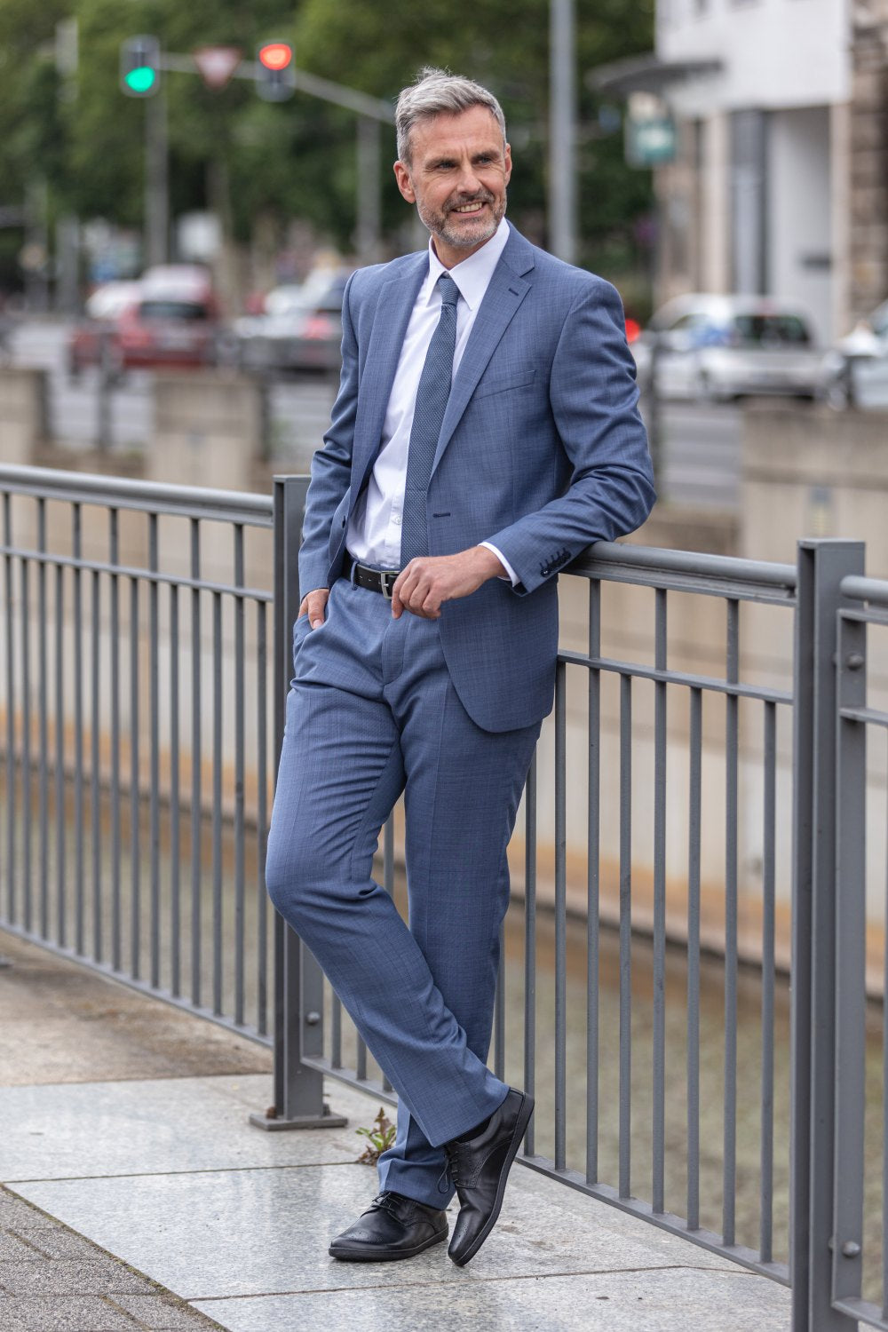 Ein Mann im blauen Anzug lehnt lässig an einem Geländer auf einer Stadtstraße und trägt klassische BRIQ Brogue Black-Schuhe von ZAQQ. Er hat graues Haar und einen Bart und hat die Hände in den Taschen. Im Hintergrund sind Autos und eine von Bäumen gesäumte Straße zu sehen.