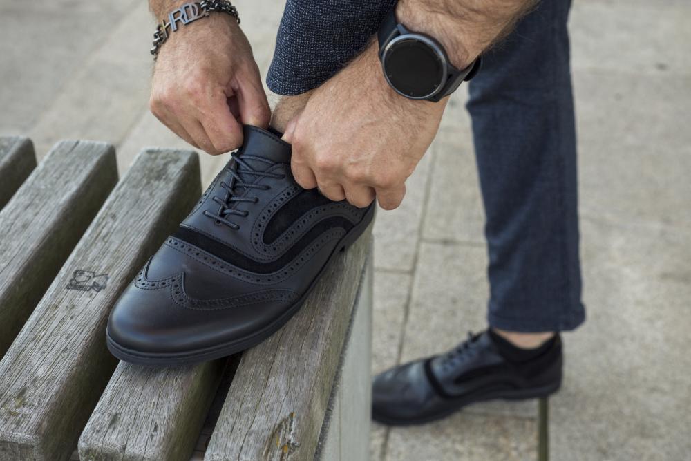 Eine Person schnürt die Schnürsenkel eines BRIQ Brogue Deep Black von ZAQQ, gefertigt aus luxuriösem Kalbsleder mit dekorativen Perforationen. Sie trägt eine Uhr und ein Armband und steht mit einem Fuß auf einer Holzbank.