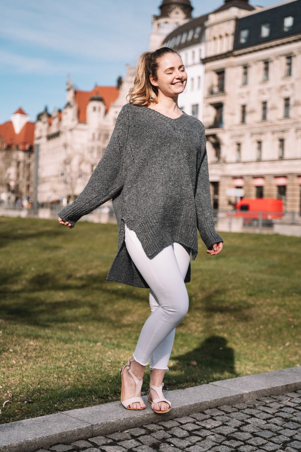 Eine lächelnde Person im Freien, die einen grauen Pullover und weiße Hosen trägt, steht in stylischen ZAQQ CLIQ Nude-Sandalen auf einem Steinweg. Im Hintergrund sieht man üppiges Gras, ein Gebäude mit rotem Dach und einen klaren blauen Himmel.