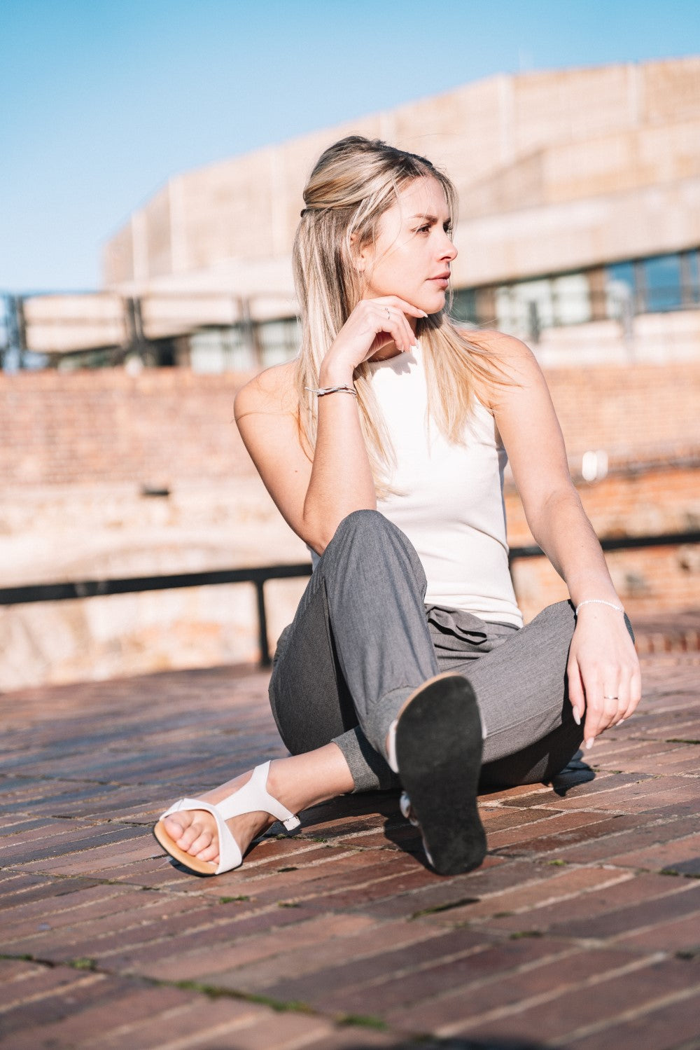 Eine Frau mit langen blonden Haaren, einem weißen ärmellosen Oberteil und grauen Hosen, sitzt auf einem Ziegelsteinpflaster und trägt die ZAQQ CLIQ White Barfuß-Sandale mit flexibler Sohle aus Nappaleder. Sie stützt ihr Kinn auf ihre Hand und blickt nachdenklich in die Ferne vor dem Hintergrund eines Ziegelsteingebäudes und des klaren blauen Himmels.