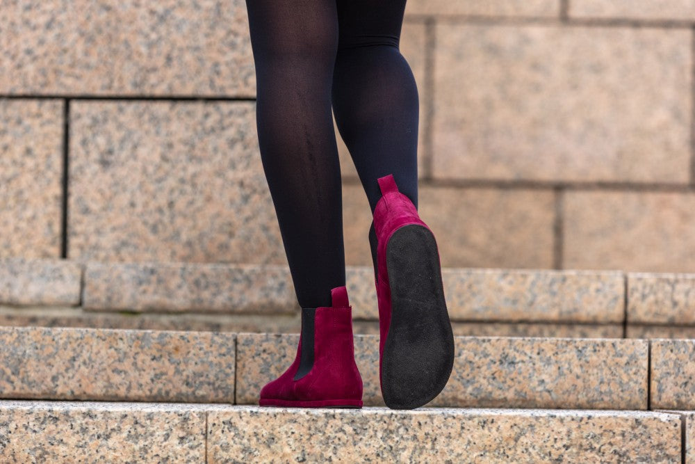 Eine Person trägt schwarze Leggings und auffällige EQUITY Berry-Boots von ZAQQ mit einem auffälligen roten Wildlederdesign und geht Steinstufen hinauf. Die Betonung des Barfußschuh-Designs unterstreicht sowohl Bewegung als auch Stil.