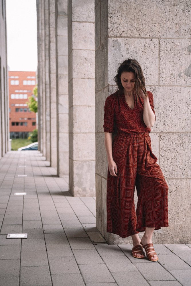 Eine Frau lehnt an einer Steinsäule in einem Außengehweg und blickt sanft nach unten. Sie trägt den FLAQE Brown Jumpsuit von ZAQQ, kombiniert mit minimalistischen Sandalen. Gebäude bilden eine entfernte Kulisse und rahmen ihren ruhigen Moment mit zeitloser Eleganz ein.