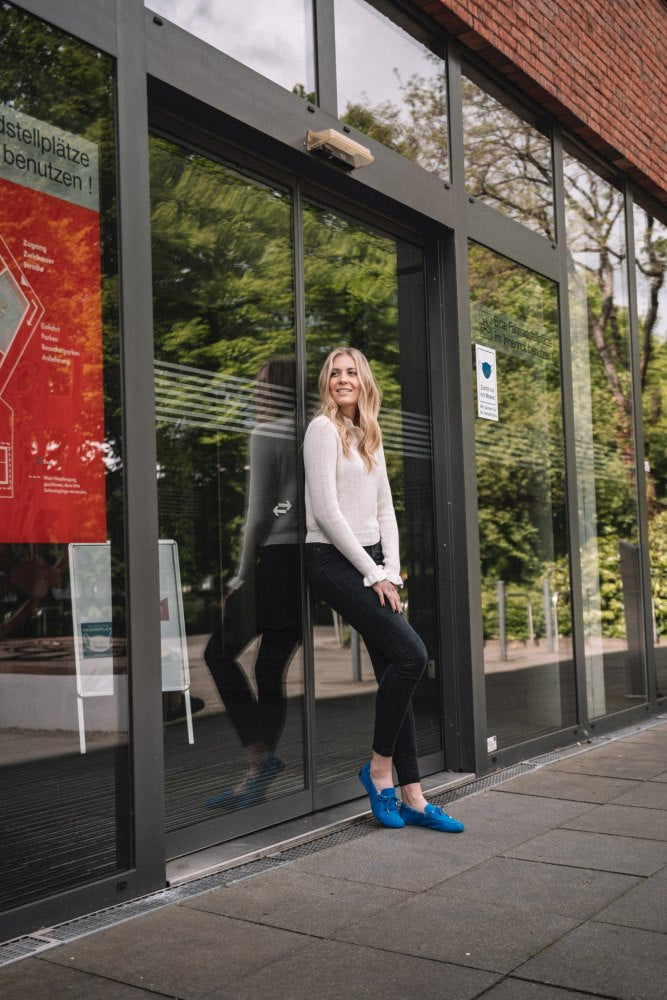 Eine Person mit langen Haaren, bekleidet mit einem hellen Pullover und dunkler Hose sowie den schicken FLIQ Blue Schuhen von ZAQQ, sitzt auf der Kante einer Glastür. Im Hintergrund schimmern Spiegelungen im Glas, neben einem roten Informationsplakat und kräftigem Grün.