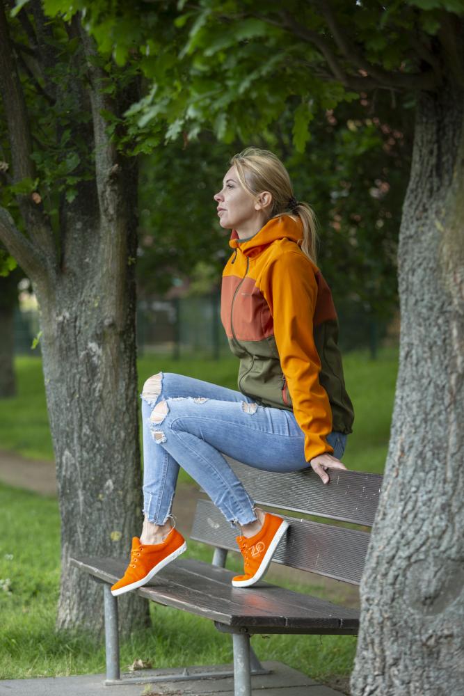 Eine Person in orange-grüner Jacke und zerrissenen Jeans sitzt auf einer Parkbank. Sie trägt leuchtende LIQE Orange Sneaker von ZAQQ, die für ein Barfußgefühl konzipiert sind, und scheint zwischen den grünen Bäumen nachdenklich in die Ferne zu blicken.