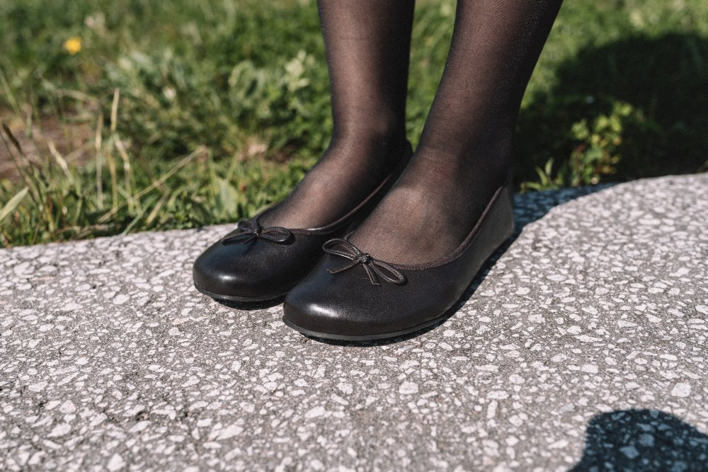 Nahaufnahme einer Person in dunkelbraunen Ballerinas und Strumpfhosen, die auf einer grauen, strukturierten Oberfläche mit Gras im Hintergrund steht – ein perfekter Moment, um mit dem LOOQ Dark Brown-Stil von ZAQQ von klassisch zu zeitgenössisch zu wechseln.