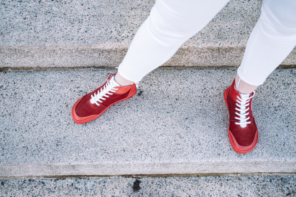 Eine Person, die LUQQ Red-Sneakers mit weißen Schnürsenkeln und weißen Hosen von ZAQQ trägt, steht auf einer grauen Steintreppe. Das von oben aufgenommene Bild hebt die flexible Kautschuksohle und den breiten Zehenbereich des Schuhs hervor und betont Komfort und Stil.