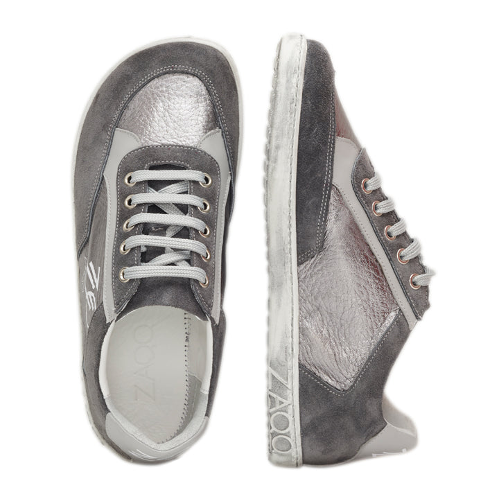 Ein Paar stylische ZAQQ OQTAV Grey Sneaker mit Veloursleder und silbermetallischen Einsätzen. Ein Sneaker ist seitlich zu sehen, während der andere von oben präsentiert wird, was die weißen Schnürsenkel und die strukturierten Gummisohlen hervorhebt.