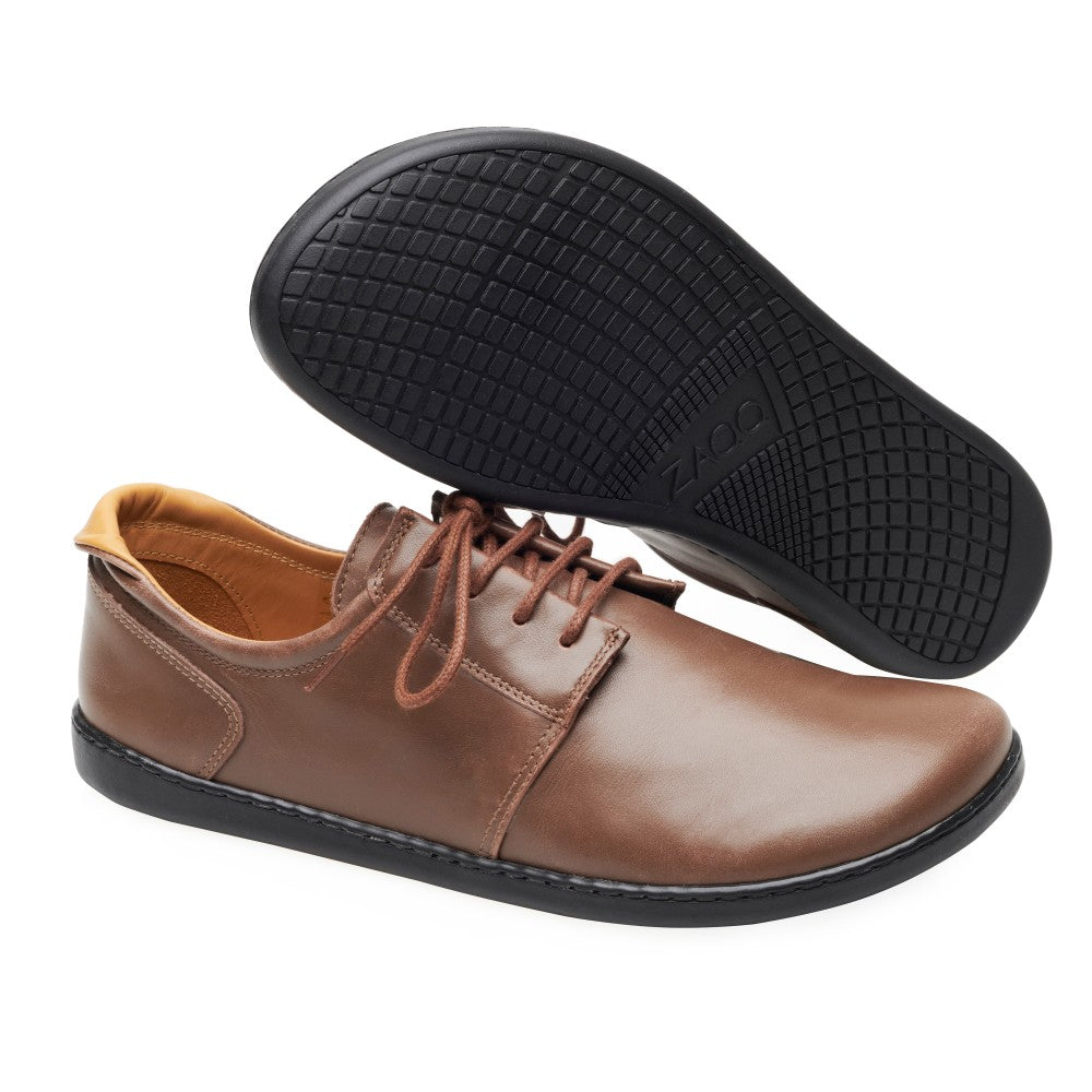 Die PIQUANT Nappa Dark Brown Schuhe von ZAQQ zeichnen sich durch ein elegantes und schlichtes Design aus. Ein Schuh steht aufrecht und betont das Seitenprofil, während der andere kopfüber steht und das schwarze strukturierte Sohlenmuster zur Geltung bringt.