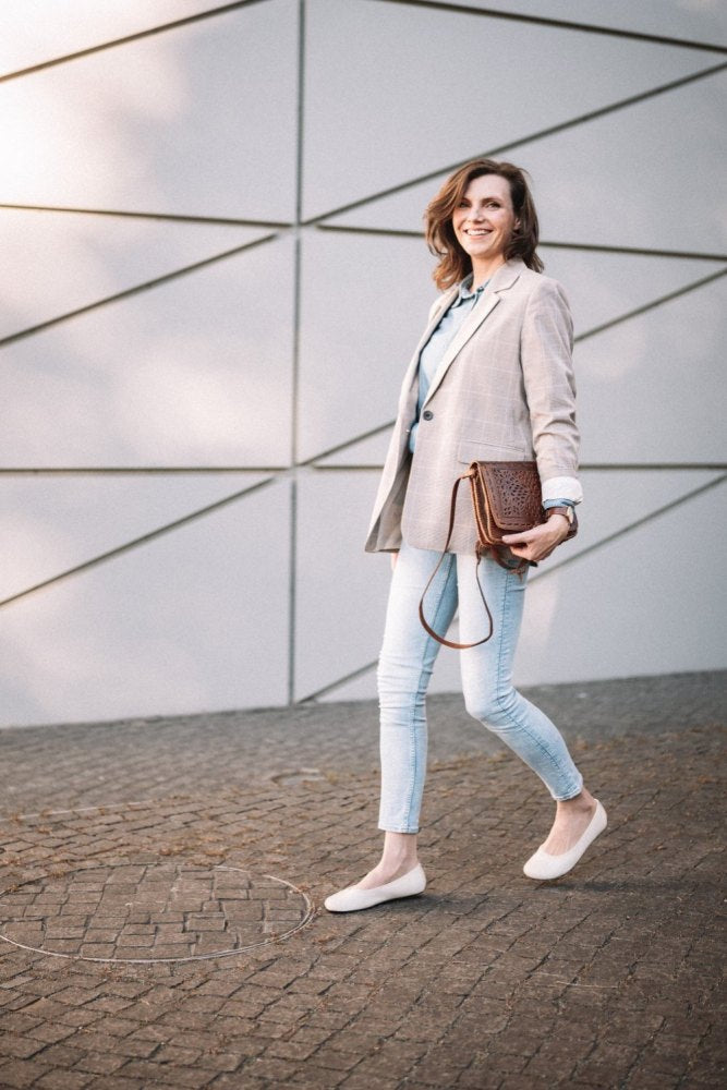 Eine Frau in einem hellkarierten Blazer und Jeans schlendert selbstbewusst in ihren beigen ZAQQ QARMA-Schuhen über einen Kopfsteinpflasterweg. Sie hält eine braune Handtasche in der Hand und lächelt vor der modernen geometrischen Wand, während ihr Barfußschuh leicht über die sonnenbeschienenen Steine tritt.