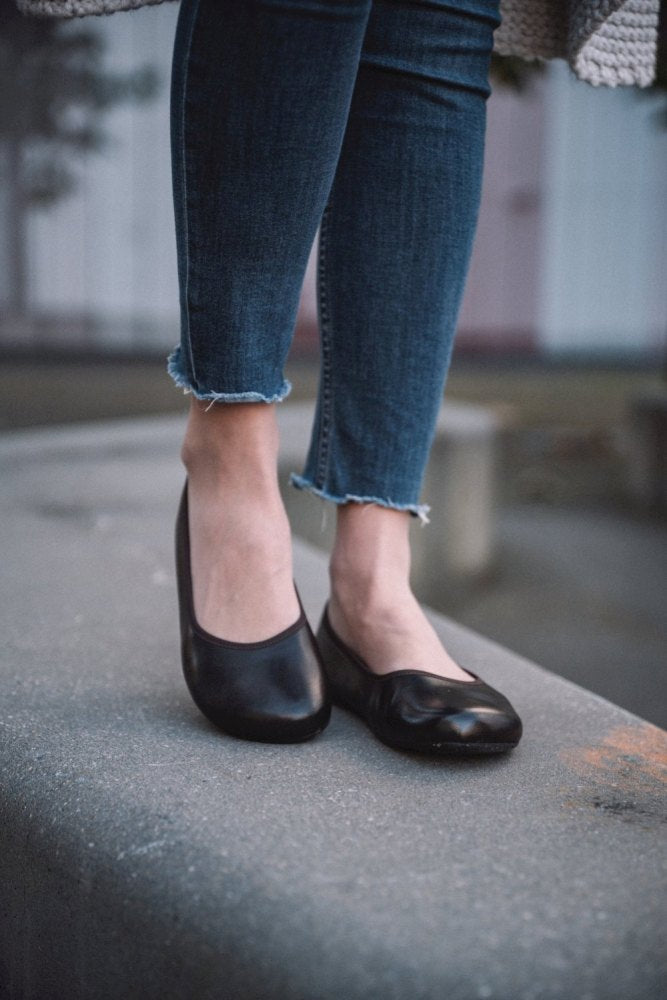 Eine Person steht auf einer Betonfläche und trägt dunkelbraune Ballerinas von ZAQQ QARMA, kombiniert mit Bluejeans mit ausgefransten Kanten. Der Hintergrund ist leicht verschwommen.