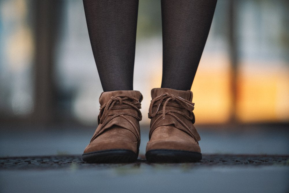 Eine Nahaufnahme einer Person, die schwarze Strumpfhosen und die Stiefeletten QEBEQ Cognac Veloursleder von ZAQQ trägt und auf einer gemusterten Oberfläche steht. Der Hintergrund weist verschwommene warme Töne auf.