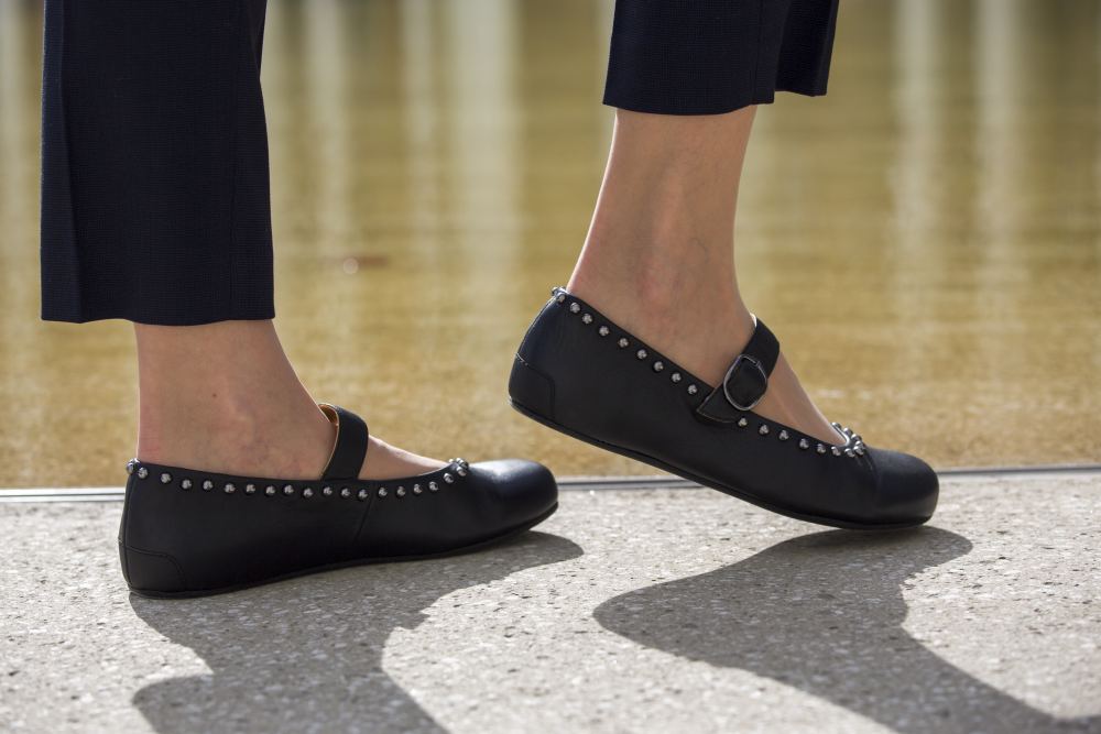 Eine Nahaufnahme zeigt eine Person, die auf eine Betonoberfläche tritt. Sie trägt die mit Nieten besetzten Ballerinas DIEQO Nappa Black von ZAQQ, die aus luxuriösem Nappaleder gefertigt und mit einem eleganten Riemen versehen sind. Die Person trägt eine schwarze Hose und die reflektierende Wasseroberfläche im Hintergrund unterstreicht die anmutige Eleganz jeder natürlichen Fußbewegung.