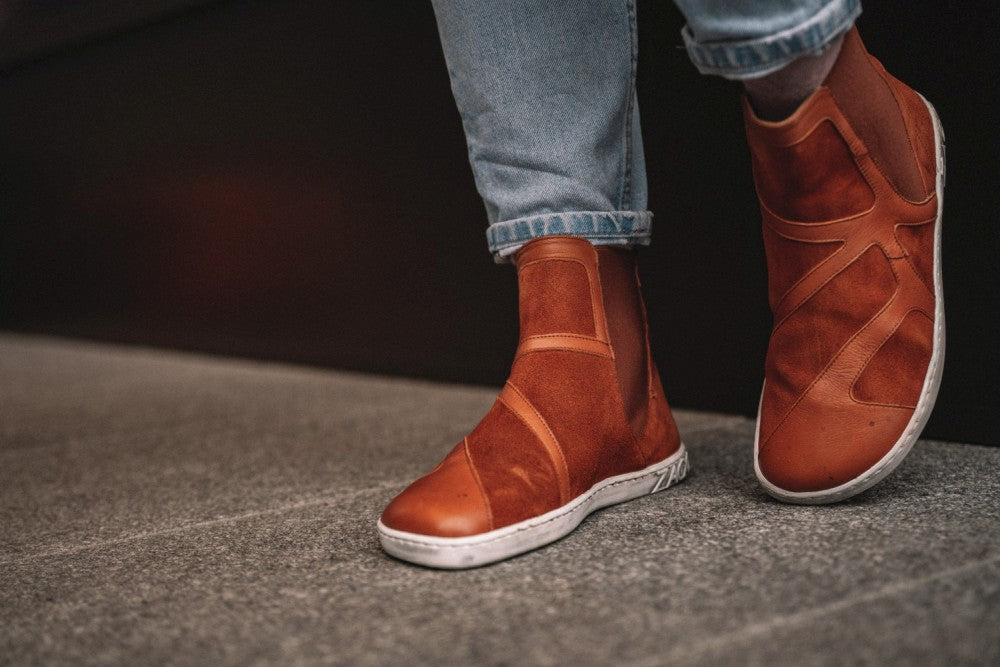 Eine Person trägt die trendigen QEY WEST Cognac High-Top-Sneaker von ZAQQ mit einem markanten geometrischen Muster und kontrastierenden weißen Sohlen. Sie steht auf einer Betonfläche und trägt hochgekrempelte Bluejeans.