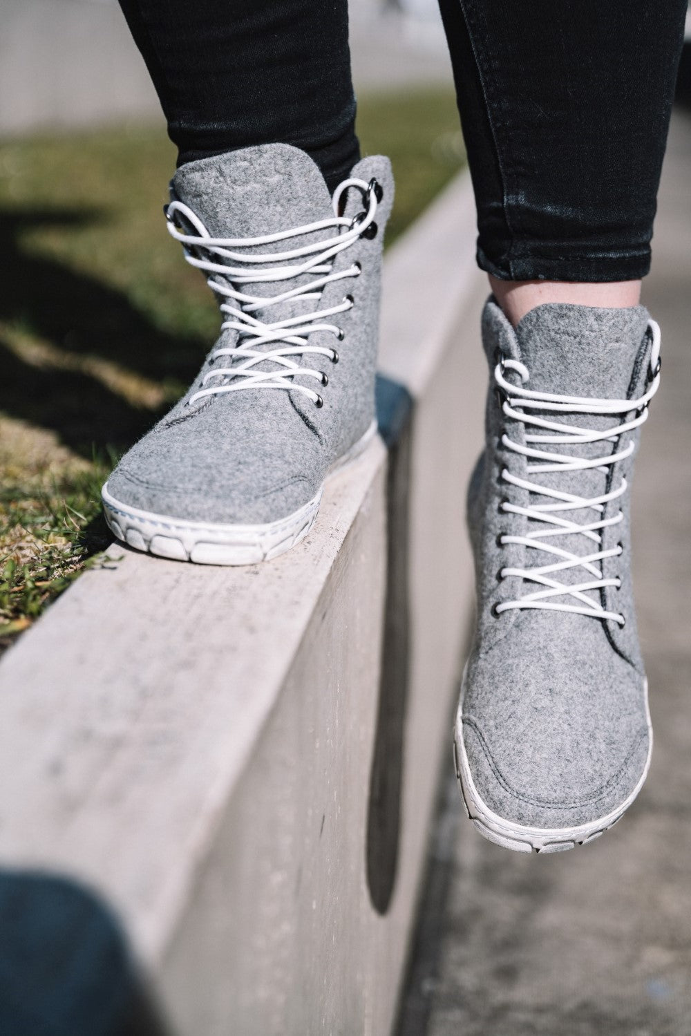 Zwei Füße in ZAQQs QICE Grey High-Top-Sneakern mit weißen Schnürsenkeln balancieren auf einer niedrigen Betonmauer und bieten mit ihrer Outdoorsohle Halt. Die Person trägt eine schwarze Hose und im Hintergrund ist eine verschwommene Ansicht von Gras zu sehen.