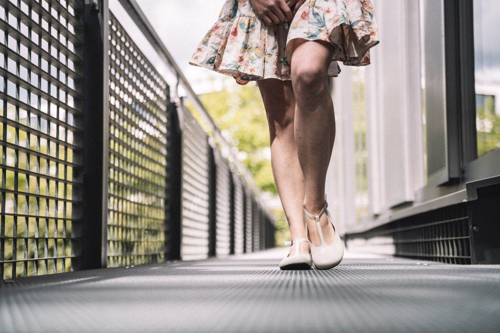 Nahaufnahme einer Frau in einem geblümten Kleid und weißen Schuhen, die über eine Metallbrücke läuft und den Komfort von ZAQQs QITE Beige zeigt. Der Fokus liegt auf ihren Beinen und Füßen vor einem unscharfen Hintergrund aus Geländer und Grün.