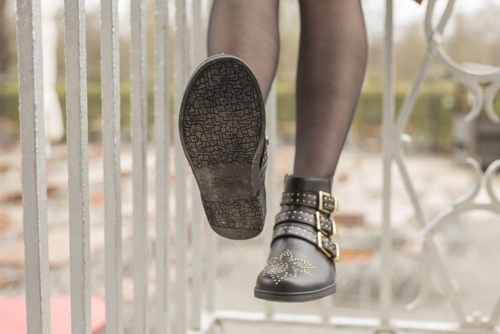 Eine Person trägt ZAQQs QLANCE Black Gold-Stiefeletten mit glänzenden Nieten und Schnallen und schreitet anmutig eine Metalltreppe hinauf. Die Sohle weist ein strukturiertes Muster auf, während der Hintergrund unscharf ist, um den Fokus auf die Eleganz und den Stil des Schuhwerks zu lenken.