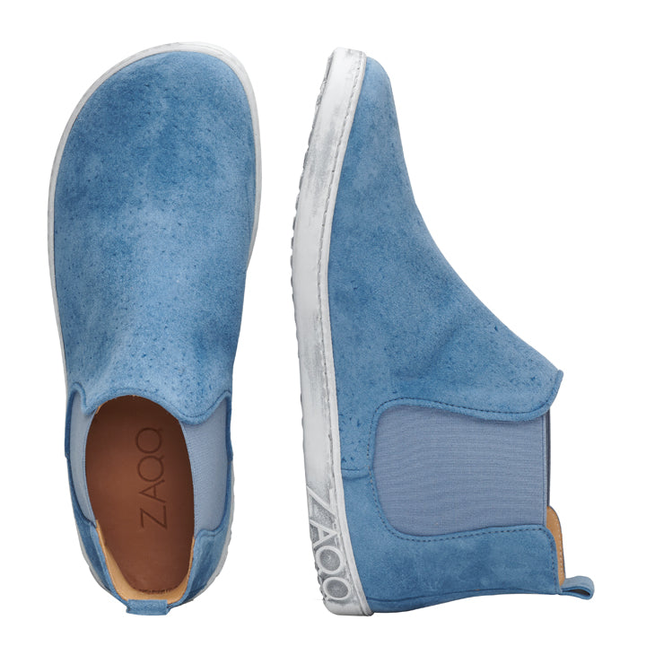 Ein Paar QOLLINS Blue Slipper von ZAQQ aus Veloursleder mit weißer Laufsohle und elastischen Seiteneinsätzen. Ein Schuh steht aufrecht, während der andere auf der Seite liegt, sodass die braune Innensohle mit dem deutlich sichtbaren Markennamen ZAQQ sichtbar ist.