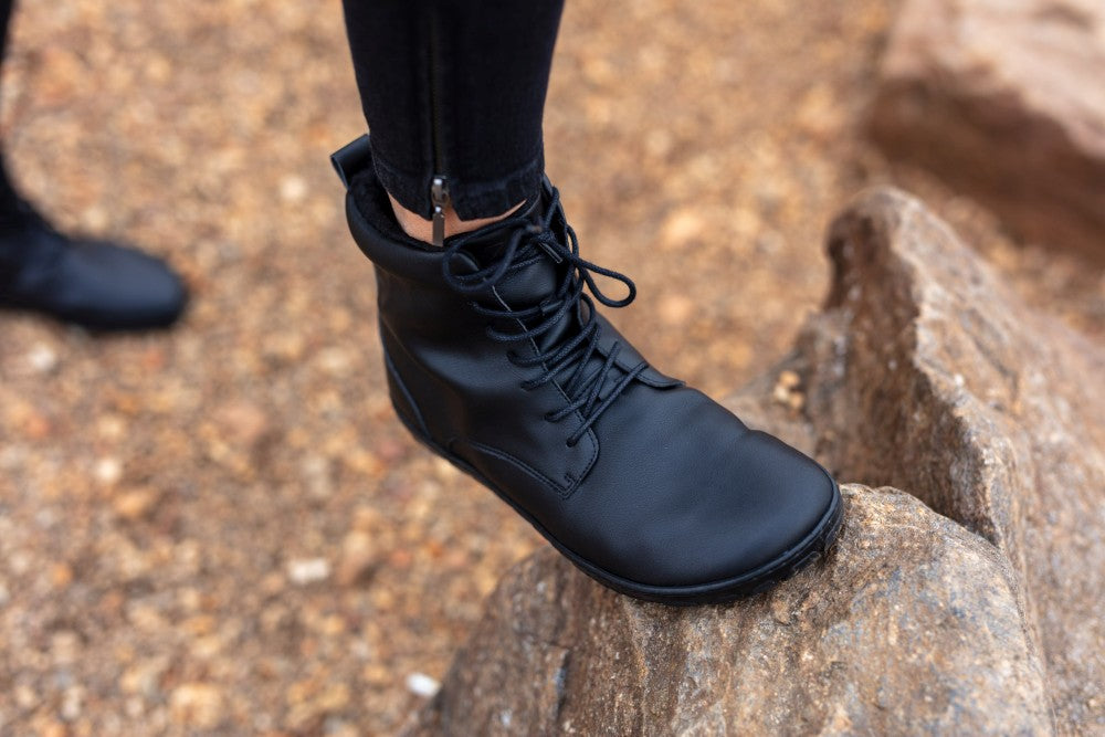 Eine Person, die die QUINTIC Winter Vegan Black-Stiefel von ZAQQ trägt, steht auf einem strukturierten Felsen, was eine Outdoor- oder raue Umgebung mit Kies im Hintergrund und einem teilweise sichtbaren weiteren Stiefel suggeriert. Diese minimalistischen Stiefel ergänzen die schwarze Hose mit Knöchelreißverschlüssen perfekt.