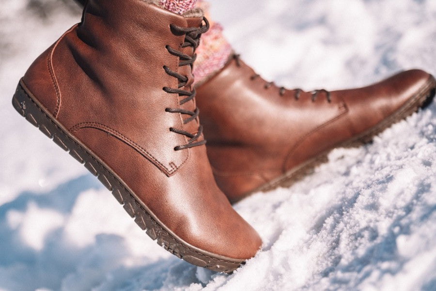 Nahaufnahme der geschnürten QUINTIC Winter Vegan Nut-Stiefel von ZAQQ beim Gehen auf Schnee. Diese veganen Schnürboots mit robuster Sohle bieten hervorragenden Halt. Im Hintergrund ist eine schneebedeckte Oberfläche zu sehen, von der nur der untere Teil der Beine und bunte Socken zu sehen sind.