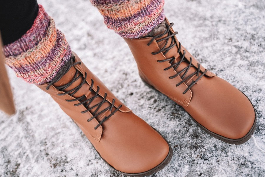 Eine Nahaufnahme zeigt eine Person, die die QUINTIC Winter Vegan Tabaco-Stiefel von ZAQQ auf einer schneebedeckten Oberfläche trägt. Die hellbraunen Stiefel haben dunkelbraune Schnürsenkel und werden stilvoll mit bunten gestrickten Beinwärmern in Rosa-, Orange- und Lilatönen kombiniert. Die Kautschuksohle bietet hervorragenden Halt, während Schneeflocken sanft den Boden bestäuben.