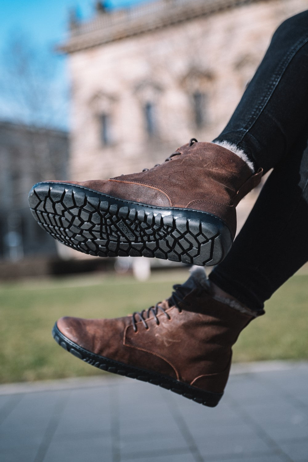 Nahaufnahme der Füße einer Person, die QUINTIC Winter Velours Brown Waterproof Wildlederstiefel von ZAQQ mit schwarzen, strukturierten Sohlen trägt. Die Stiefel heben sich leicht von einem verschwommenen Hintergrund aus Gras und Architektur ab. Der Himmel ist klar und sonnig, was auf einen angenehmen Tag im Freien schließen lässt.