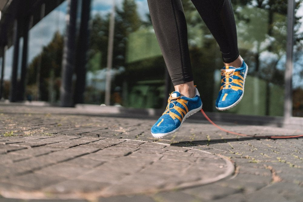 Eine Person springt Seil auf einem Ziegelsteinpflaster. Sie trägt schwarze Leggings und die ZAQQ RIQ Blue Orange Sneakers, die aus atmungsaktivem Meshgewebe bestehen. Im Hintergrund sind verschwommene Bäume und Gebäude zu sehen.