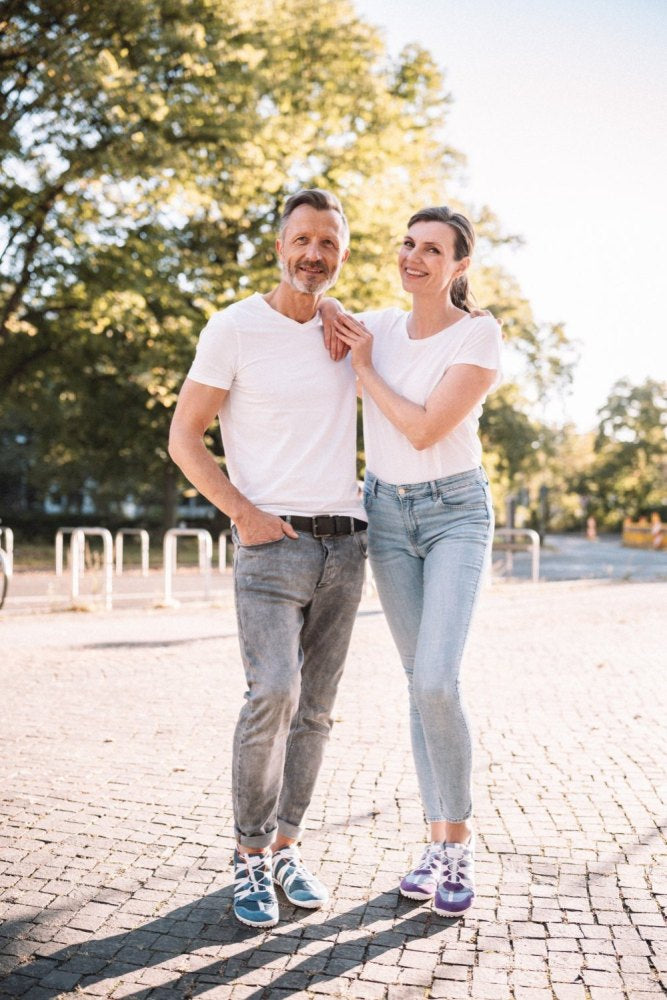 Ein Mann und eine Frau stehen lächelnd an einem sonnigen Tag im Freien. Beide tragen weiße Hemden, Jeans und die RIQ Blue-Schuhe von ZAQQ. Sie stehen auf einer gepflasterten Fläche mit Bäumen und blauem Himmel im Hintergrund.