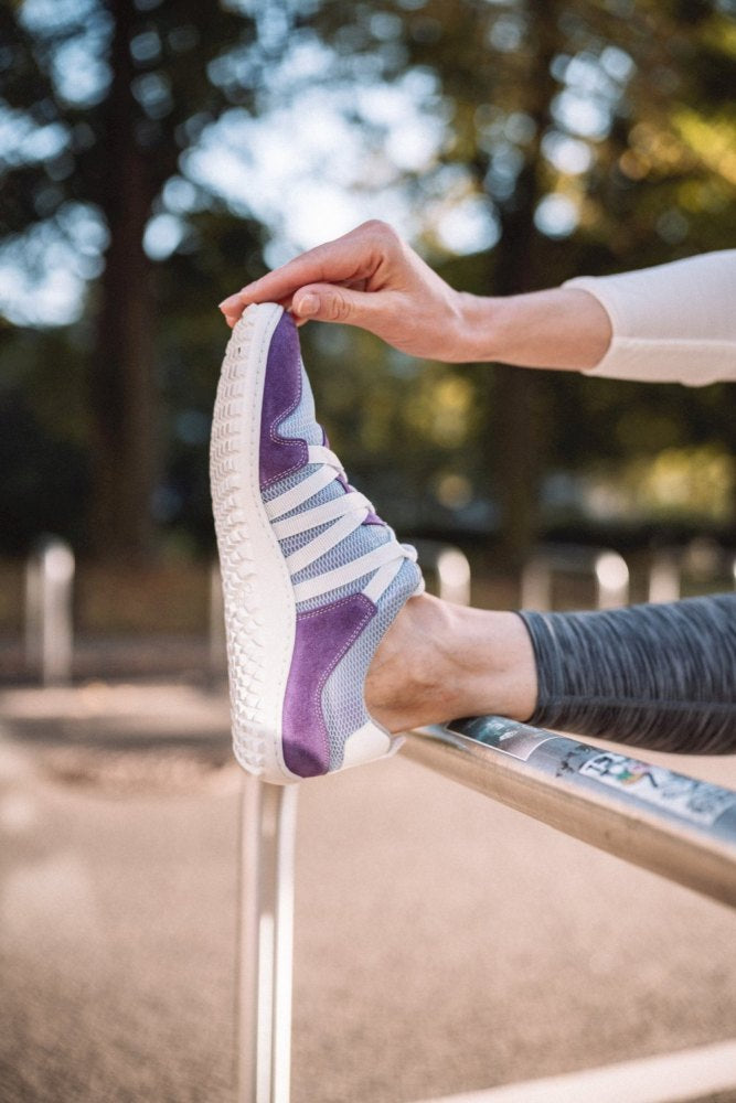 In den RIQ Purple Sneakers von ZAQQ aus atmungsaktivem Meshgewebe streckt eine Person in weißem Langarmshirt und grauer Leggings ihre Beine auf einem Geländer aus. Im Hintergrund ist eine unscharfe Parkszene mit Bäumen zu sehen.