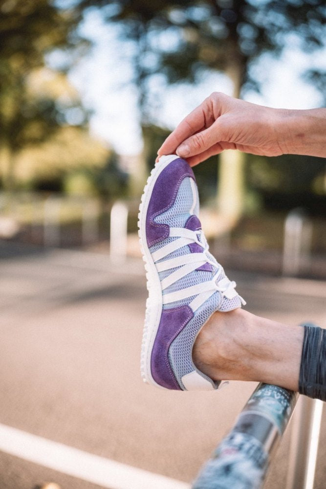 Eine Person streckt ihre Wade, wobei ein Fuß auf einem Geländer abgestützt ist. Sie trägt den RIQ Purple Sneaker von ZAQQ, der für Barfußkomfort konzipiert wurde. Die Außenkulisse zeigt Bäume und eine verschwommene Straße im Hintergrund, was die stilvolle Verwendung von Meshgewebe im Sneaker hervorhebt.