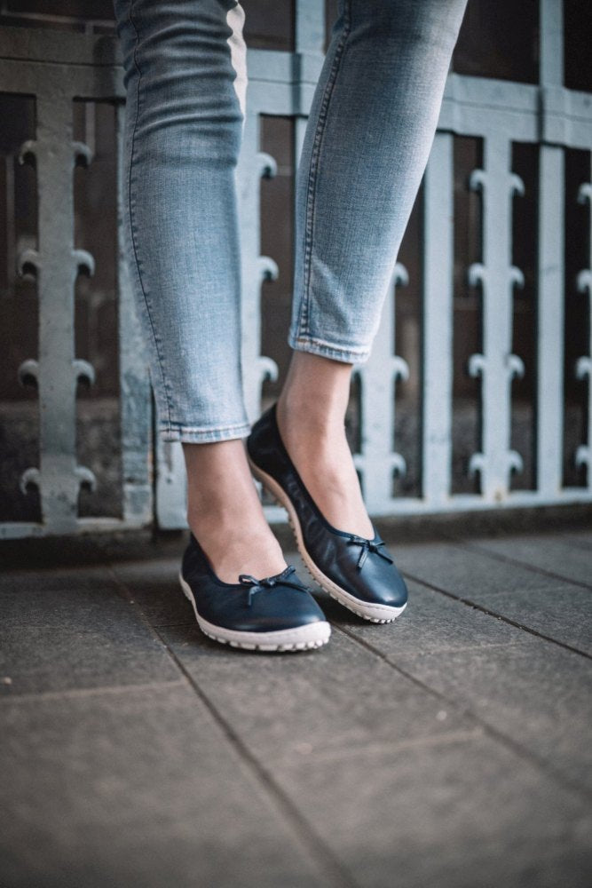 Eine Person in hellblauen Röhrenjeans steht auf einem gefliesten Untergrund und trägt ZAQQs SUQAR Darkblue, marineblaue Flats mit weißer Sohle und kleinen Schleifen, die ein dezentes Sneakersohle-Design aufweisen. Im Hintergrund ist ein hellblaues Metalltor zu sehen.