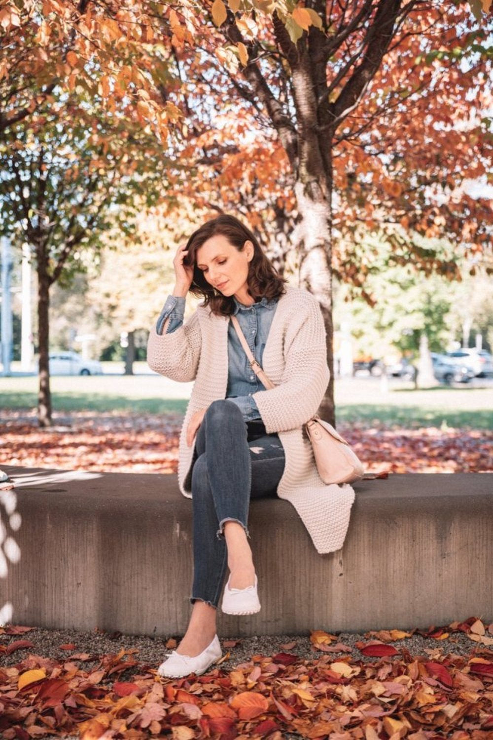 Eine Person sitzt auf einem Betonvorsprung in einem Park, umgeben von Herbstlaub. Sie trägt eine weiße Strickjacke über einem Jeanshemd und Jeans, dazu weiße SUQAR-Schuhe von ZAQQ aus Nappaleder. Bäume mit orangefarbenen Blättern bilden den Hintergrund, während Sonnenlicht durch die Zweige fällt.