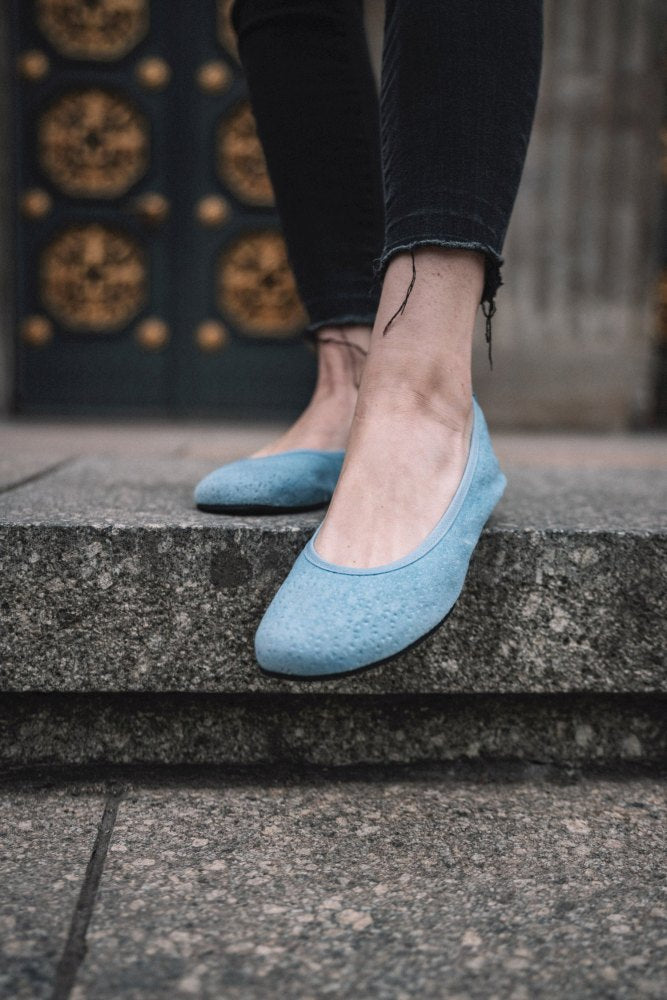 Eine Person in den TWIST Light Blue Schuhen von ZAQQ steht auf einer Steinstufe. Die schwarze Jeans der Person ist am Saum leicht ausgefranst. Im Hintergrund ist eine dekorative dunkle Tür mit goldenen Details zu sehen, die den sommerlichen Look verstärkt.