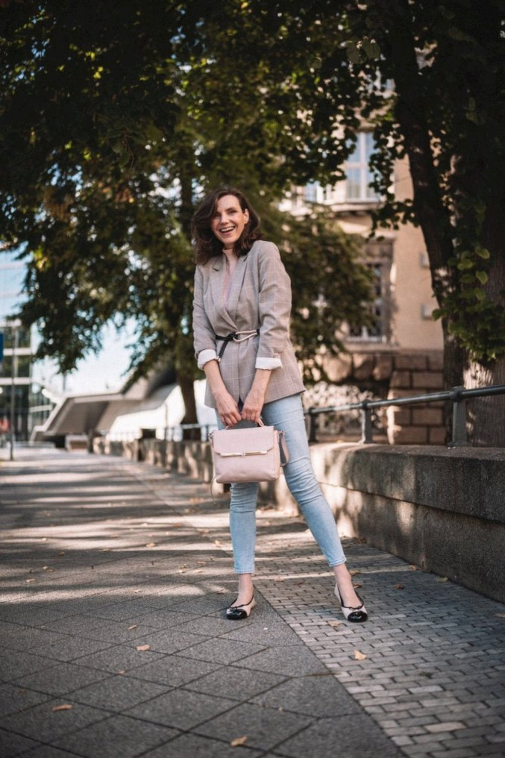 Eine Frau steht lächelnd auf einem sonnenbeschienenen Weg. Sie trägt einen hellgrauen Blazer und Jeans. Sie trägt eine beige Handtasche und trägt die ZAQQ TWIST Varnish Nude Schuhe mit Nappaleder und Kautschuksohlen, die sich perfekt in die urbane und doch natürliche Umgebung aus Bäumen und Gebäuden einfügen.